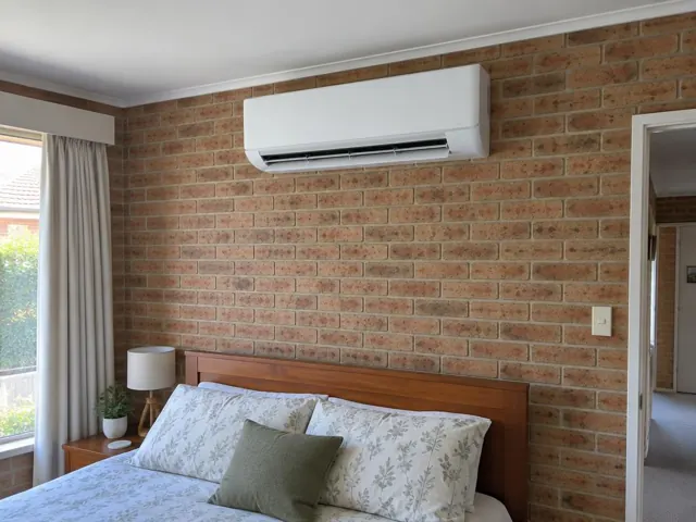 Wall-mounted split system above a bed in a brick bedroom with curtains and bedside table in Elizabeth Park